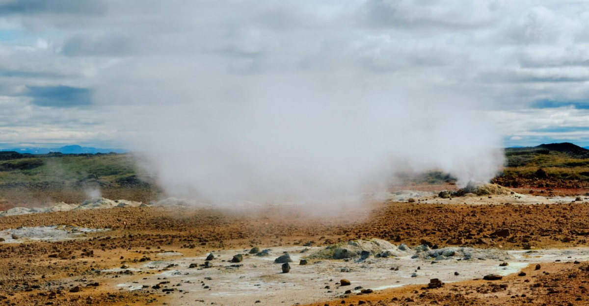 Wide view of a steaming geothermal field with cloudy sky, showcasing natural geothermal activity.