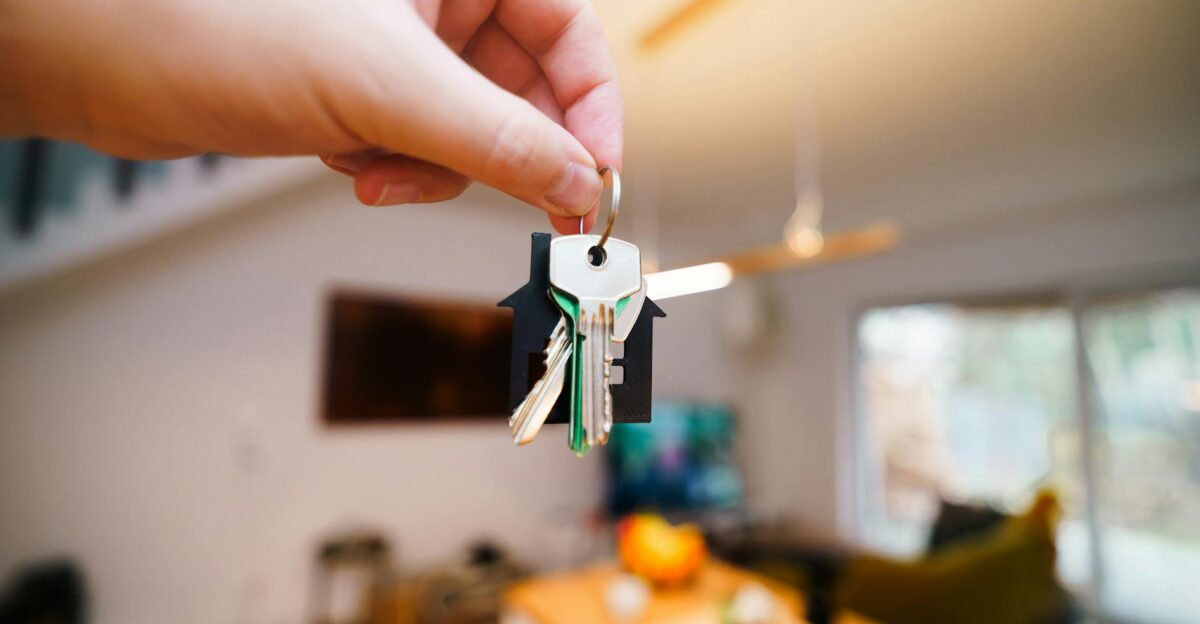 A hand holds keys with a house keychain inside a modern well-lit living space
