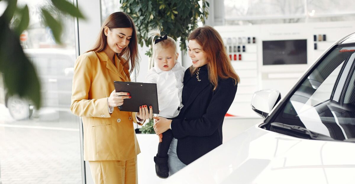 Positive young female with cute little daughter on hands discussing car characteristics with professional dealer in stylish beige suit while standing in car showroom in daylight