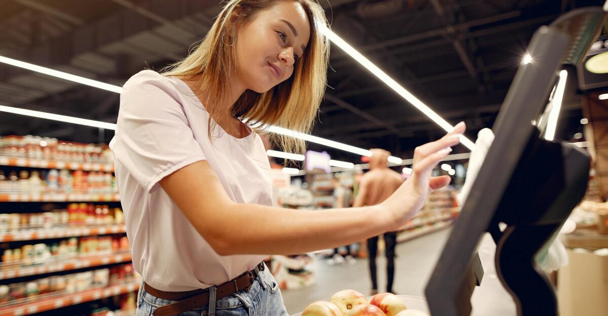 Side view of young woman in trendy clothes weighing peaches on scales while shopping in supermarket during purchase food
