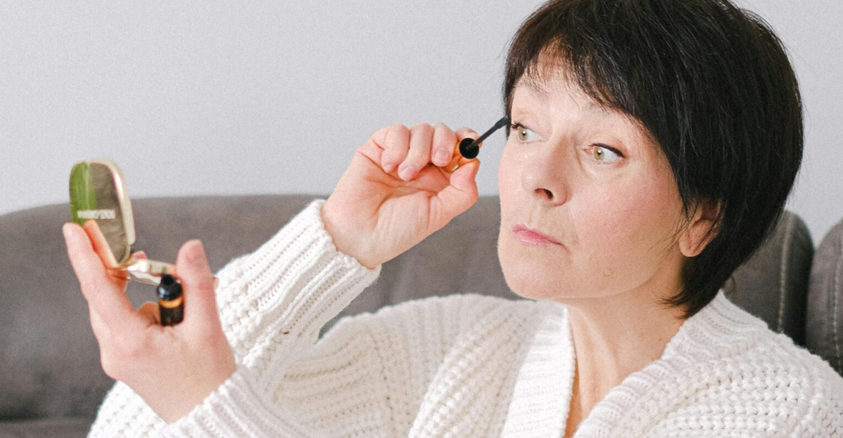 A senior caucasian woman sitting on a couch applying mascara in a cozy setting.