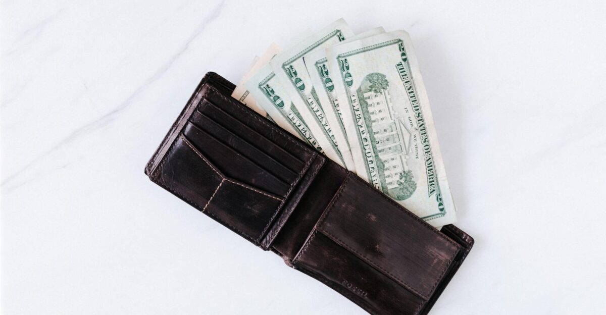 From above of black leather wallet with pockets filled with heap of dollar banknotes on white table