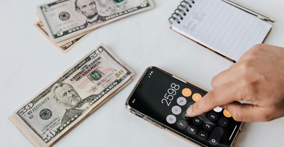 From above of crop anonymous economist calculating total amount of income using calculator app on cellphone near piles of greenbacks and notebook with pen