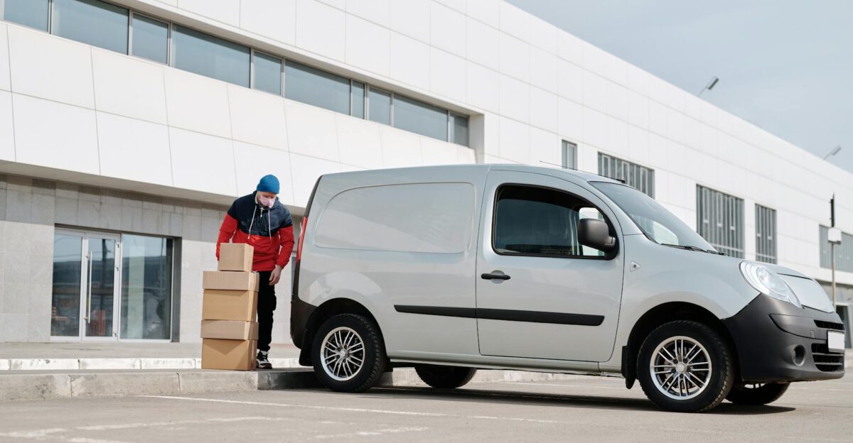 A delivery driver wearing a face mask unloads packages from a van outside a modern building