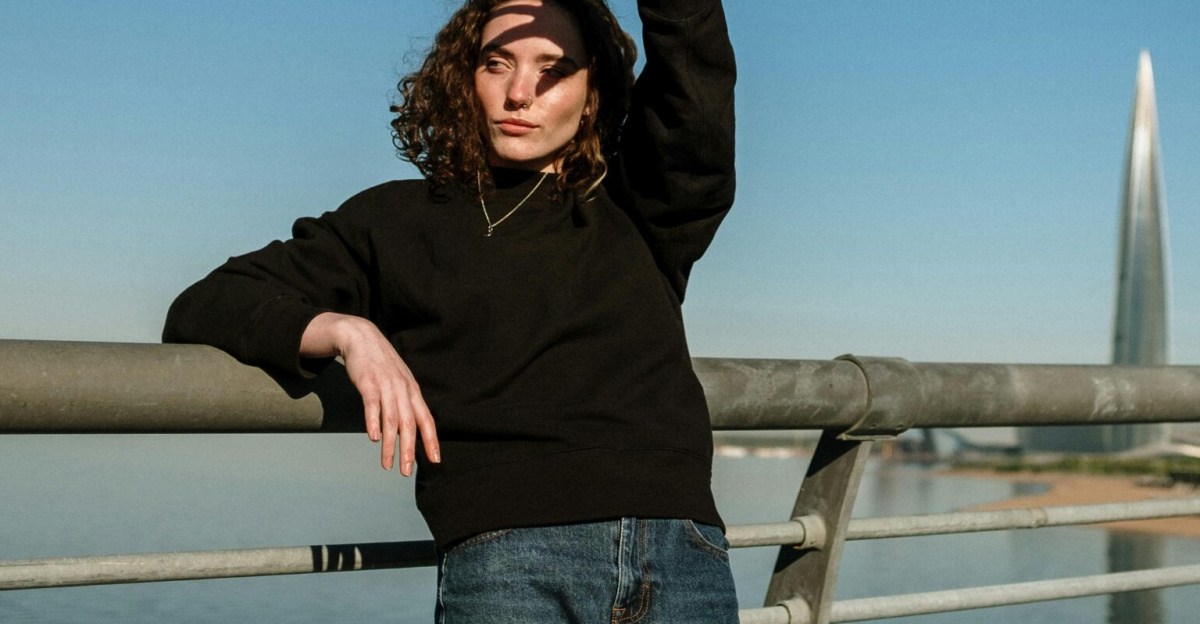 A young woman in casual attire poses on a bridge with a cityscape background featuring a modern skyscraper.