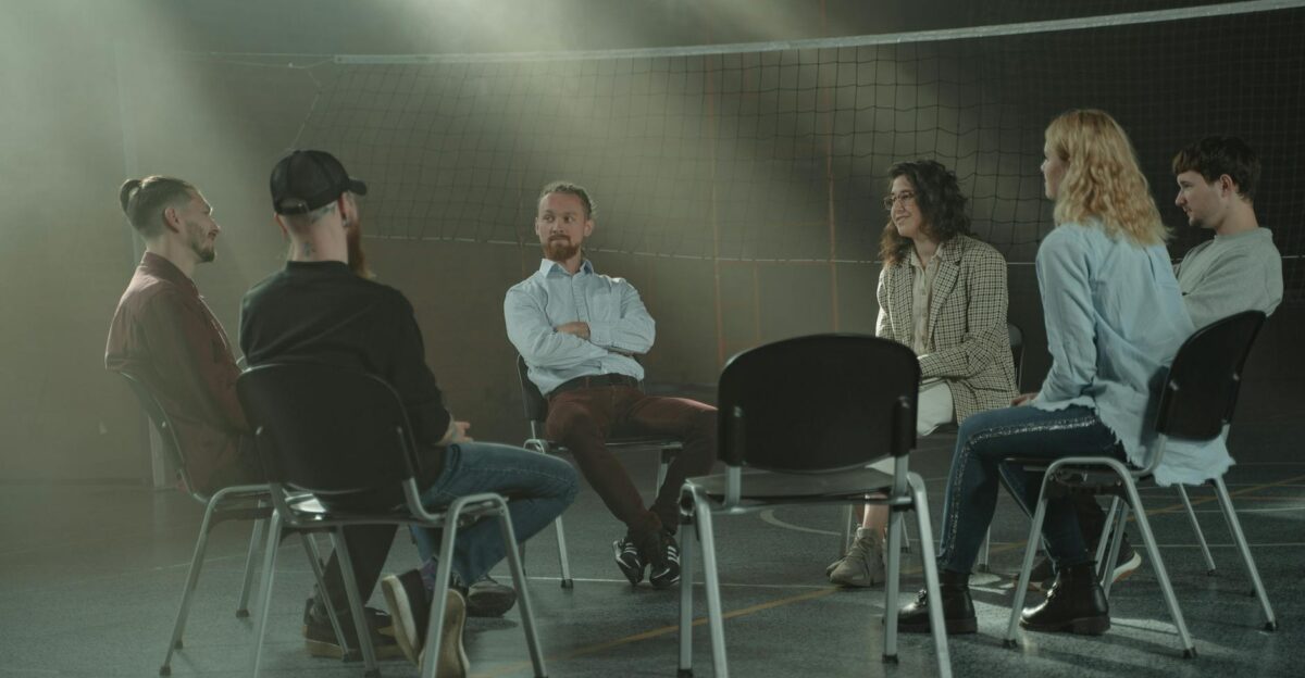 A group therapy session with six adults seated in a circle discussing support and mental health