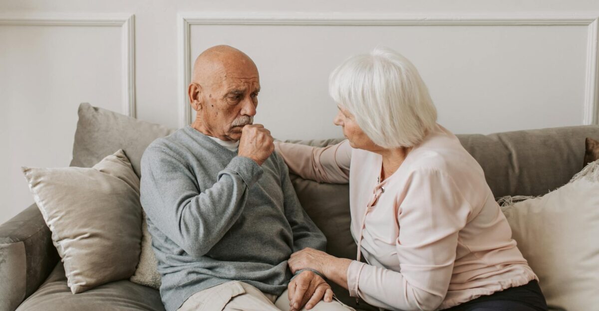 An elderly woman comforts a man coughing on a couch showcasing care and affection