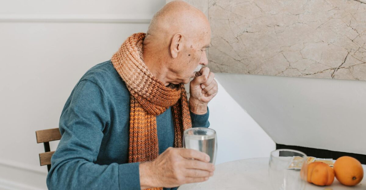 Elderly man in blue sweater coughing holding water at white table with medicines