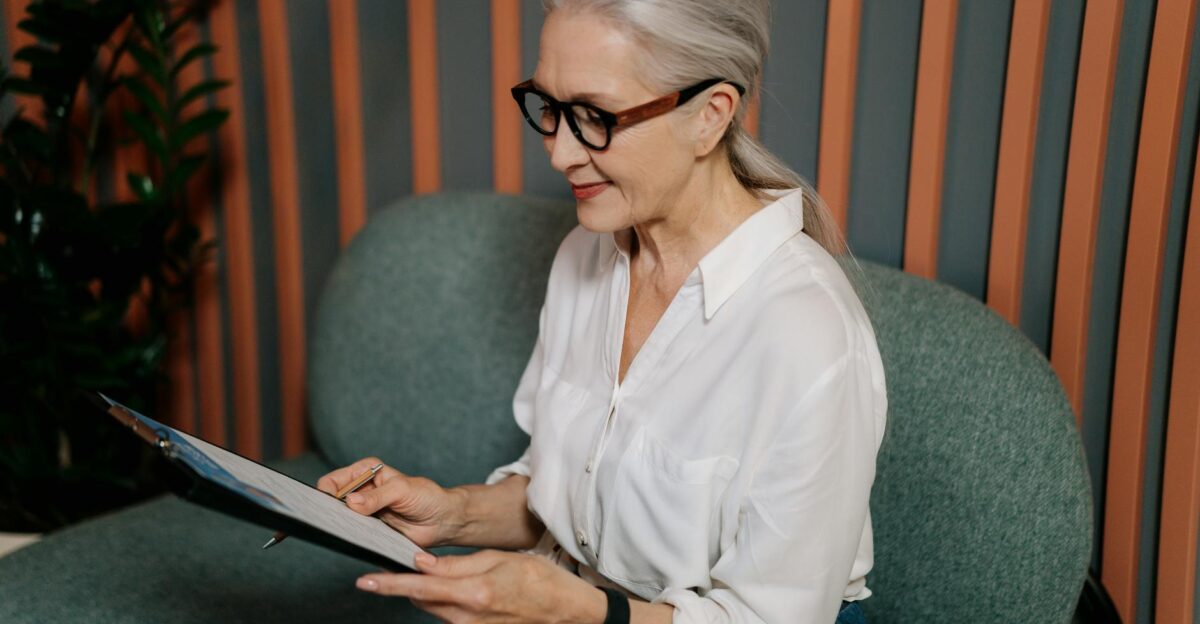 Elderly woman in glasses reviewing documents on clipboard sitting indoors with a focused expression