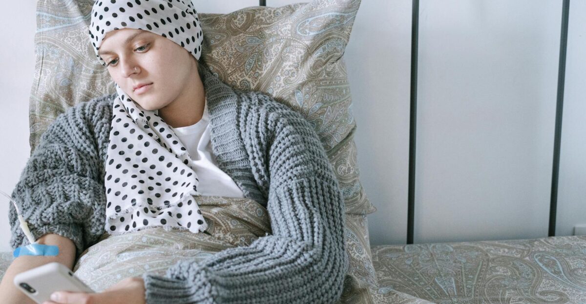 Woman resting in bed wearing a scarf using her phone during cancer treatment