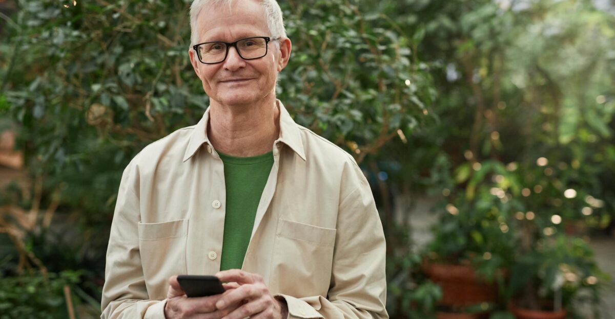 Senior man standing in a greenhouse garden smiling while using a smartphone Natural sunlight