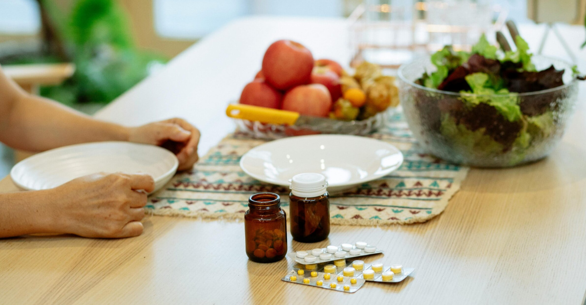 From above of crop anonymous person sitting at table with pile of various medicines and bowls of fresh lettuce salad and ripe fruits in kitchen