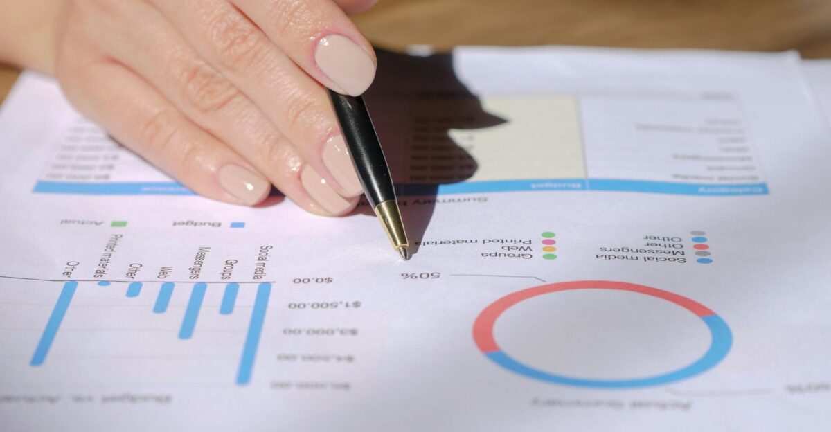 Close-up of a hand with a pen pointing at a financial document with graphs and charts