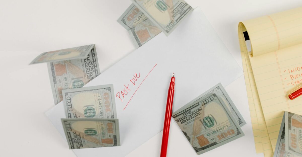A top-down view of scattered US dollar bills with a past due envelope red pen and notepad