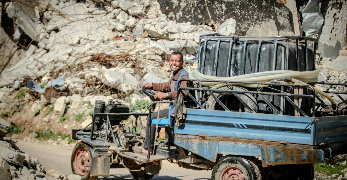 Smiling man driving a makeshift vehicle through rubble-strewn street in Idlib Syria under bright sunlight