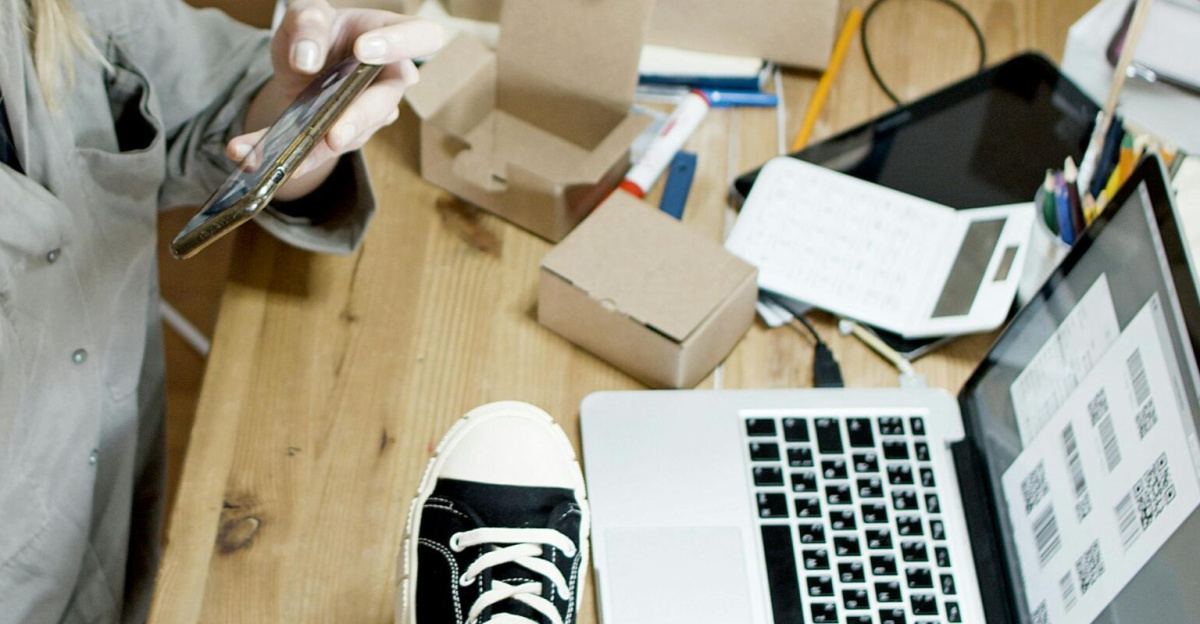 Woman photographing shoes for online sale in home workspace with laptop and packages.