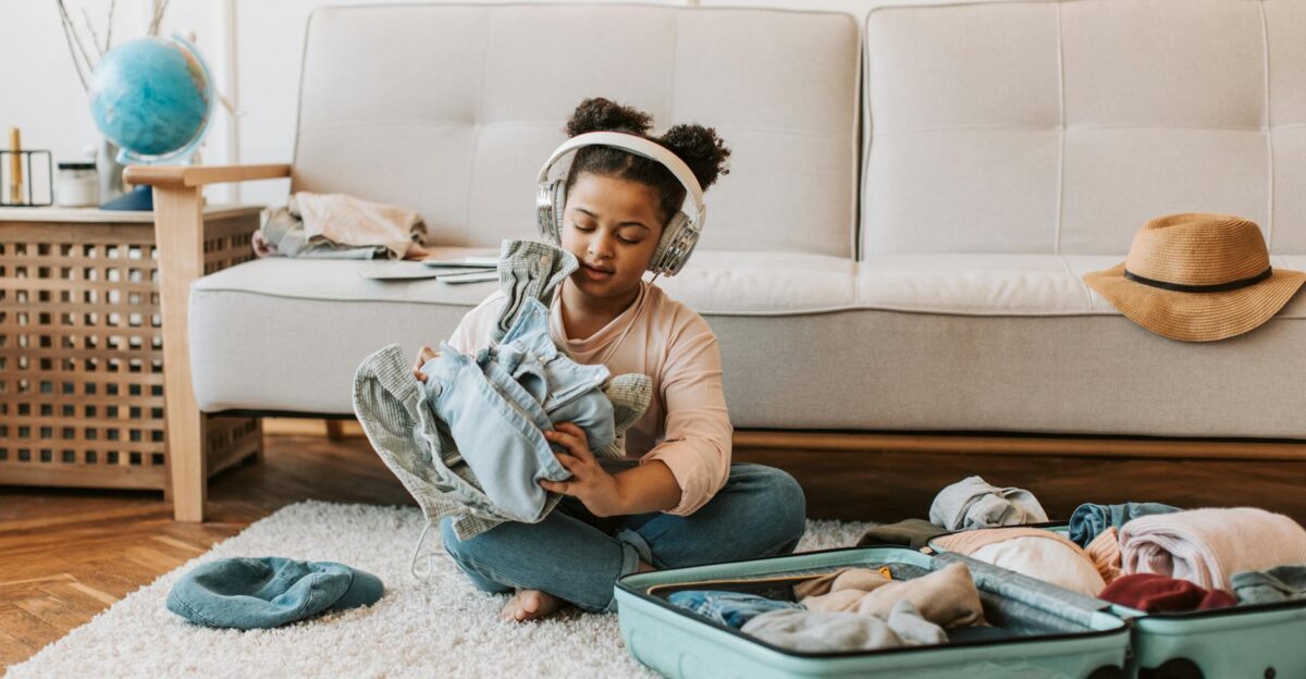 Girl sitting indoors wearing headphones folding clothes into a suitcase for a trip