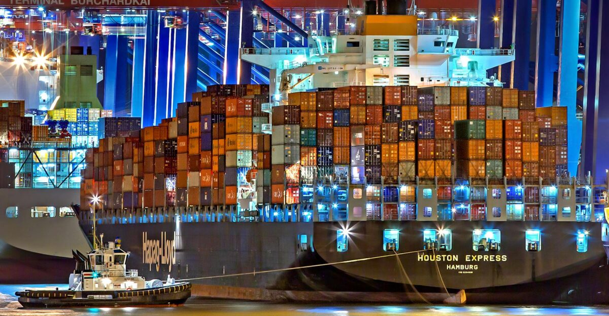 A brightly lit cargo ship at Hamburg harbor with stacked containers and a tugboat