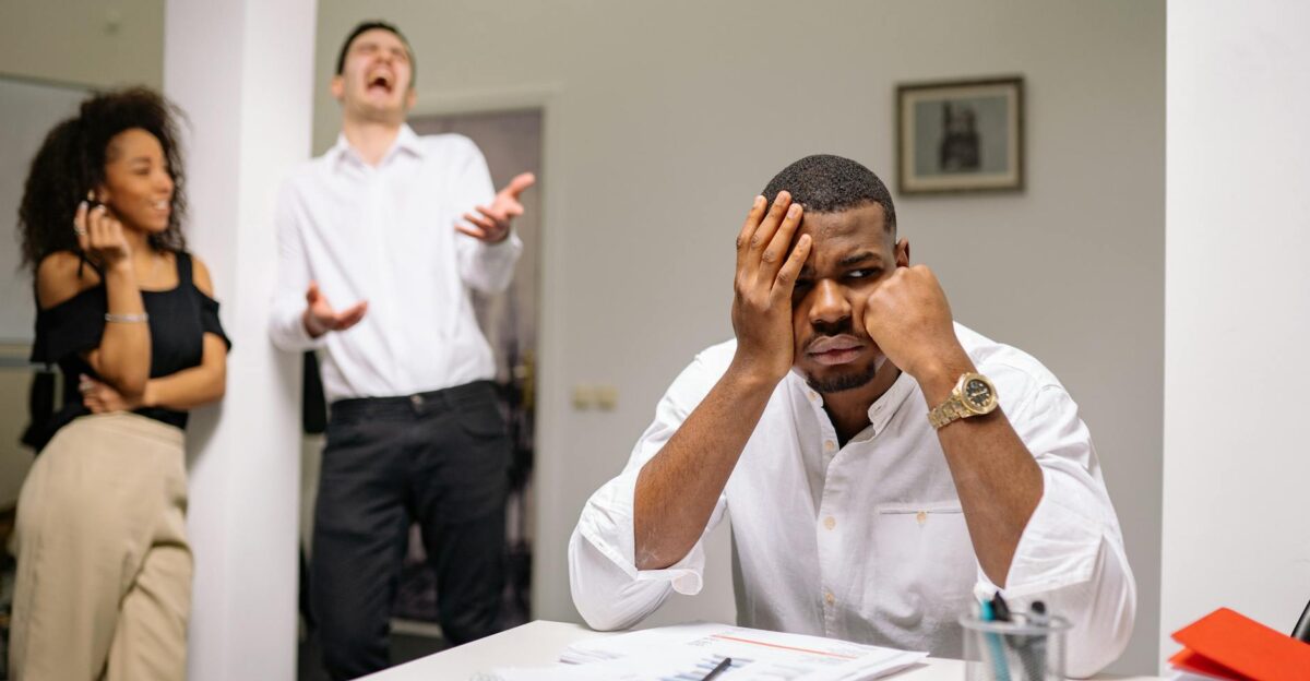 A man at a desk looks frustrated while two coworkers laugh Office workplace conflict concept