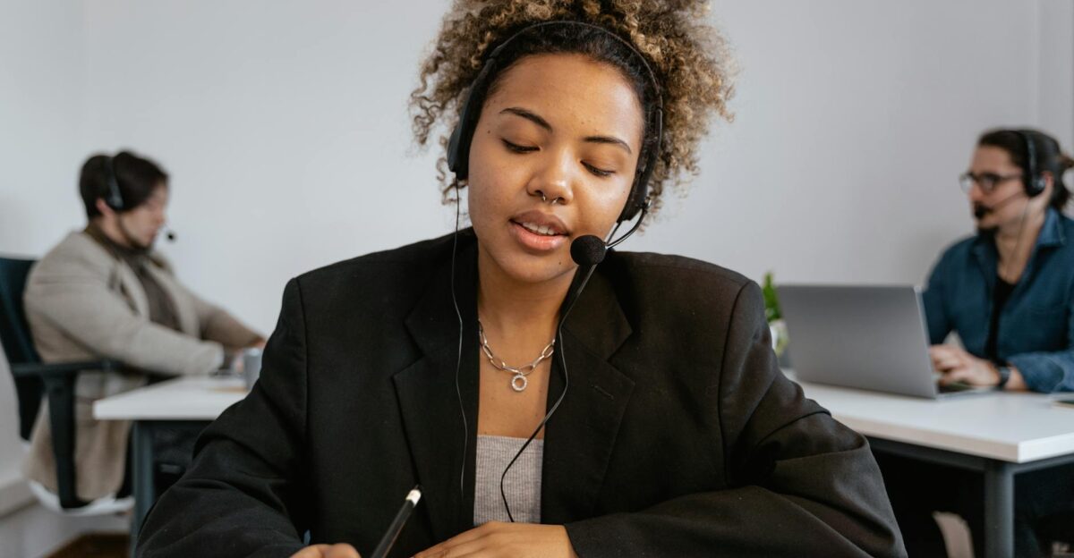 African American woman with headset working in an office with colleagues using a pen for note-taking
