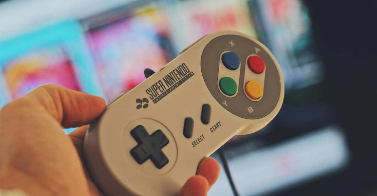 Hand holding a classic Super Nintendo game controller indoors with a blurred screen in the background
