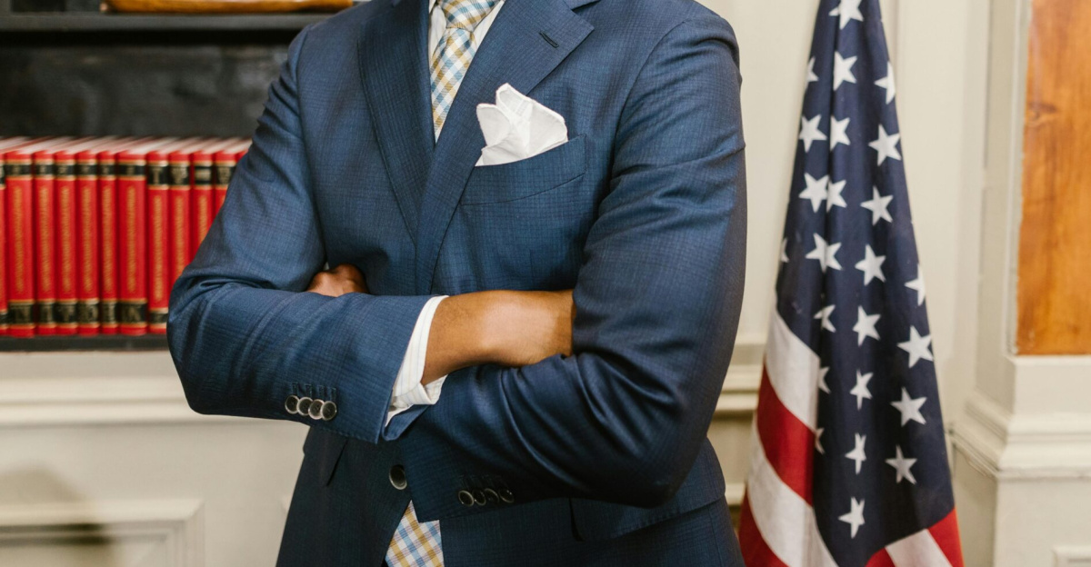 Portrait of a confident lawyer in office attire, standing with arms crossed in front of a USA flag.