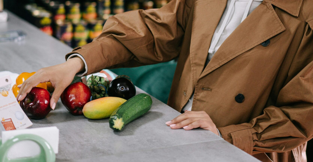 Woman with afro hair in a brown coat selecting fresh vegetables at a store counter, smiling.