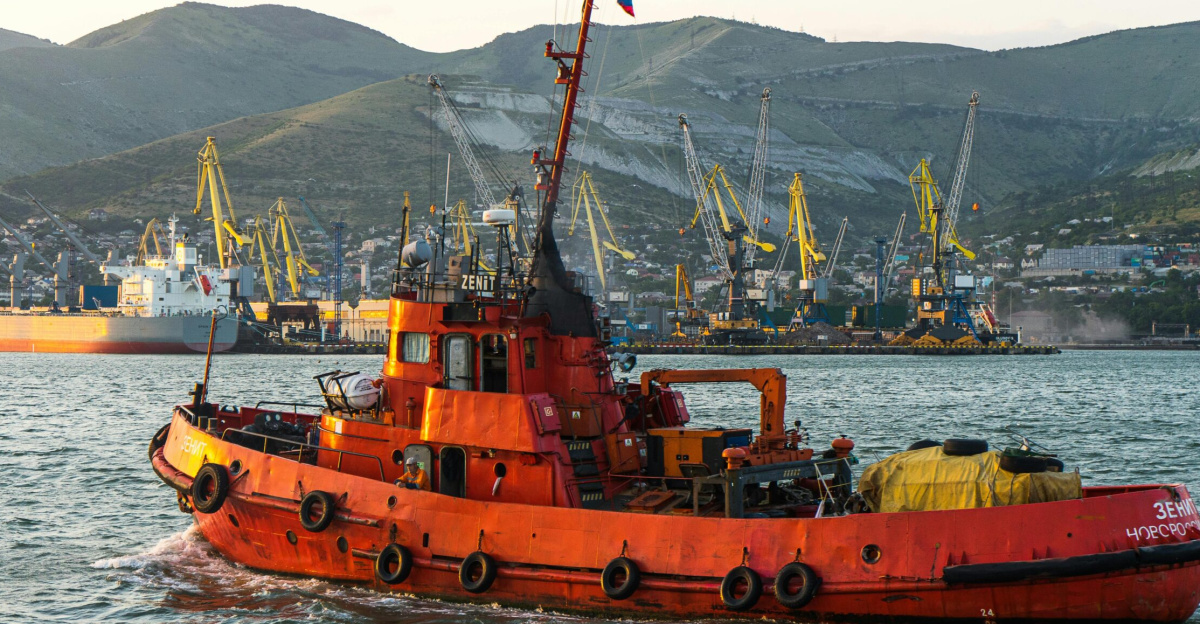 A vibrant red tugboat sails through the busy port of Novorossiysk, Russia, against scenic mountainous backdrop.
