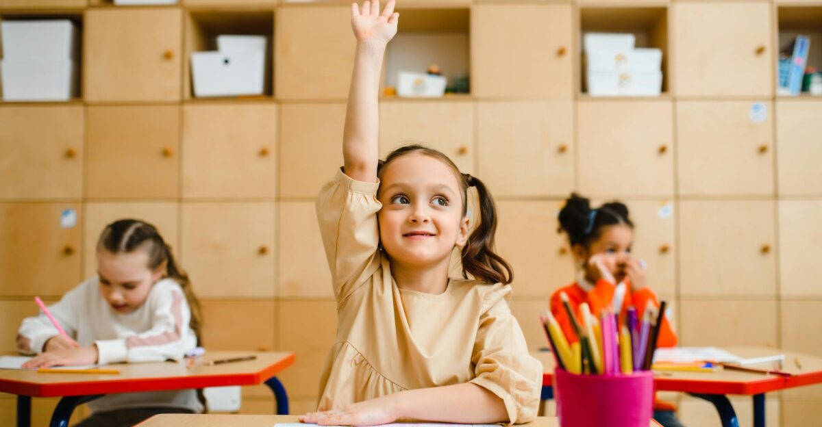 Three children engaged in learning activities inside a classroom with one raising her hand
