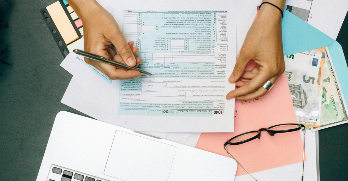 Hands writing on tax documents with laptop, glasses, and currency on desk.