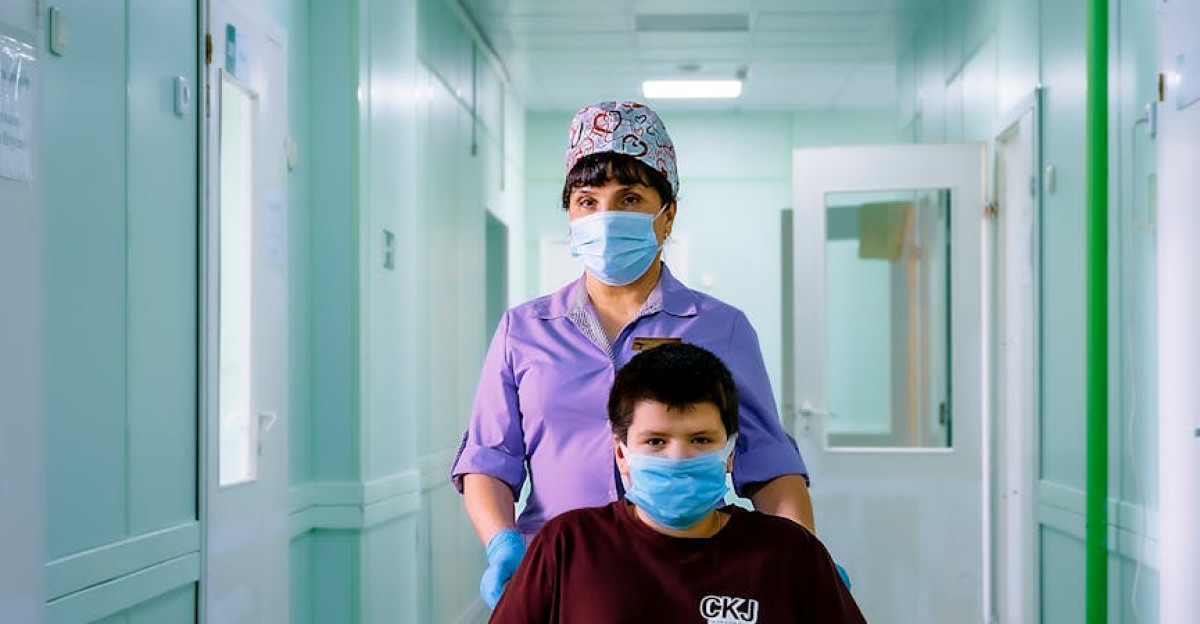 Nurse wearing mask pushes patient in wheelchair down hospital hallway