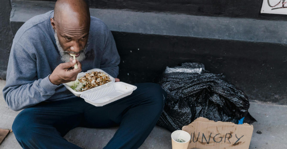 Homeless man with food container and 'hungry' sign on the sidewalk.