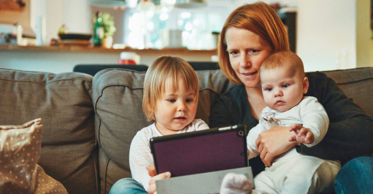two babies and woman sitting on sofa while holding baby and watching on tablet