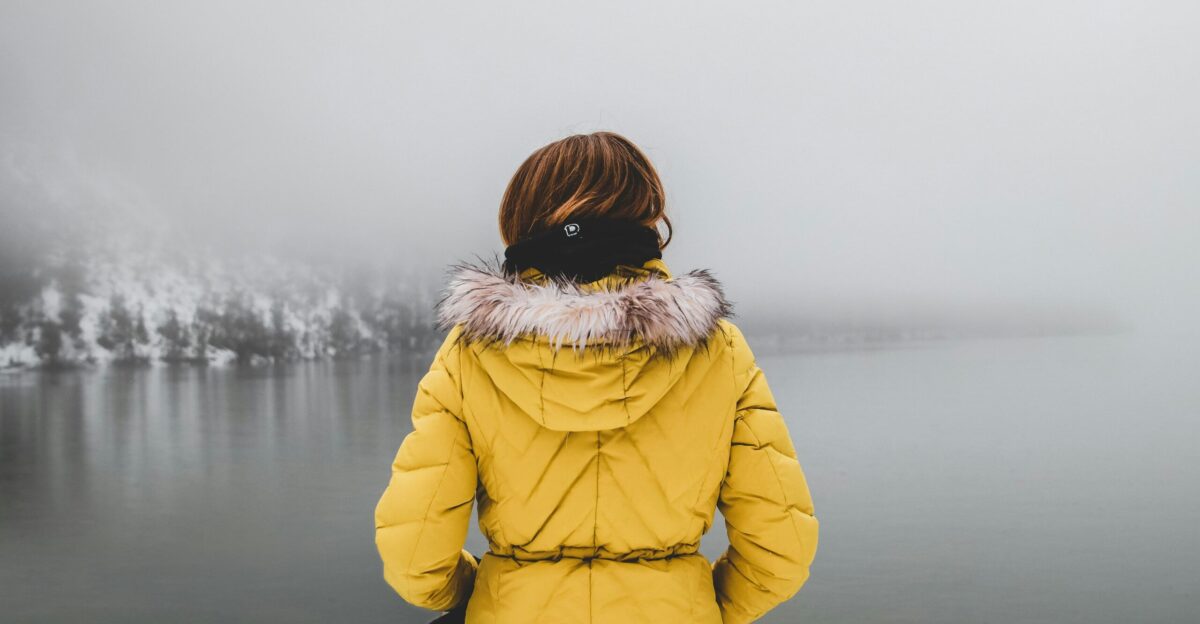 woman in yellow coat looking at body of water covered with fogs