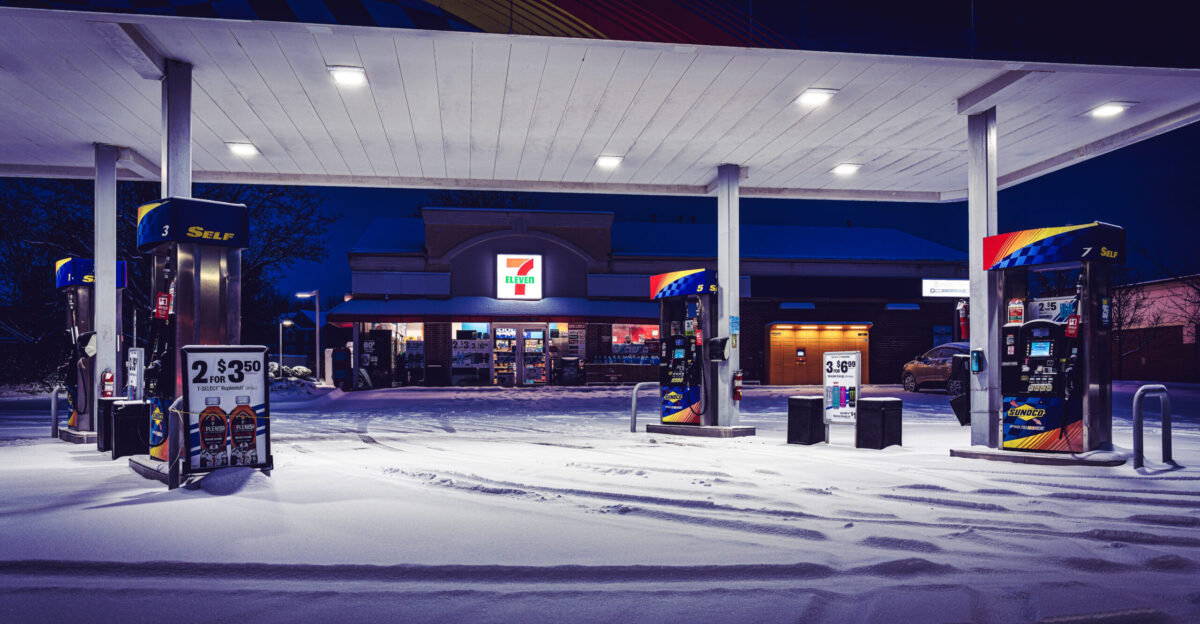 What Google Vision makes of this photo The image shows a gas station at night in the snow The foreground is dominated by a layer of freshly fallen snow showing tire tracks from cars that recently passed In the background a 7-Eleven store is brightly lit contrasting with the dark night sky The gas pumps stand under a large canopy also covered in a light dusting of snow The snow appears undisturbed around the base of the pumps suggesting that they haven t been used recently There is a car parked near the store partly visible in the background The scene is deserted there are no people visible in the image The overall mood is quiet and peaceful even somewhat desolate due to the lateness of the hour and the snowy conditions The gas station itself appears to be in a suburban or rural setting Given the apparent lack of any pedestrian activity around the well-lit 7-Eleven one might assume it is late at night and there s been minimal traffic during this time The image may have been captured from a DSLR camera considering the clarity depth of field and low-light performance likely at night based on the lighting conditions The subtle color grading in the image suggests post-processing edits in image editing software The subtle details reveal more about the scene The slight variations in the snow s texture indicate differing compaction and melting levels revealing subtle nuances of wind patterns or different foot vehicle traffic densities The price signs on the gas pumps show that the cost of fuel is 3 59 per gallon The illumination suggests the photograph was likely taken between late evening and early morning given the deep blue hues of the twilight sky There s a slight difference in lighting between the gas pumps the pumps on the left side appear to be slightly brighter than those on the right hinting at either different lighting setups or slightly different exposure during image capture The overall composition and quiet atmosphere suggest a certain degree of contemplation capturing a momentary pause in the otherwise busy life of the gas station