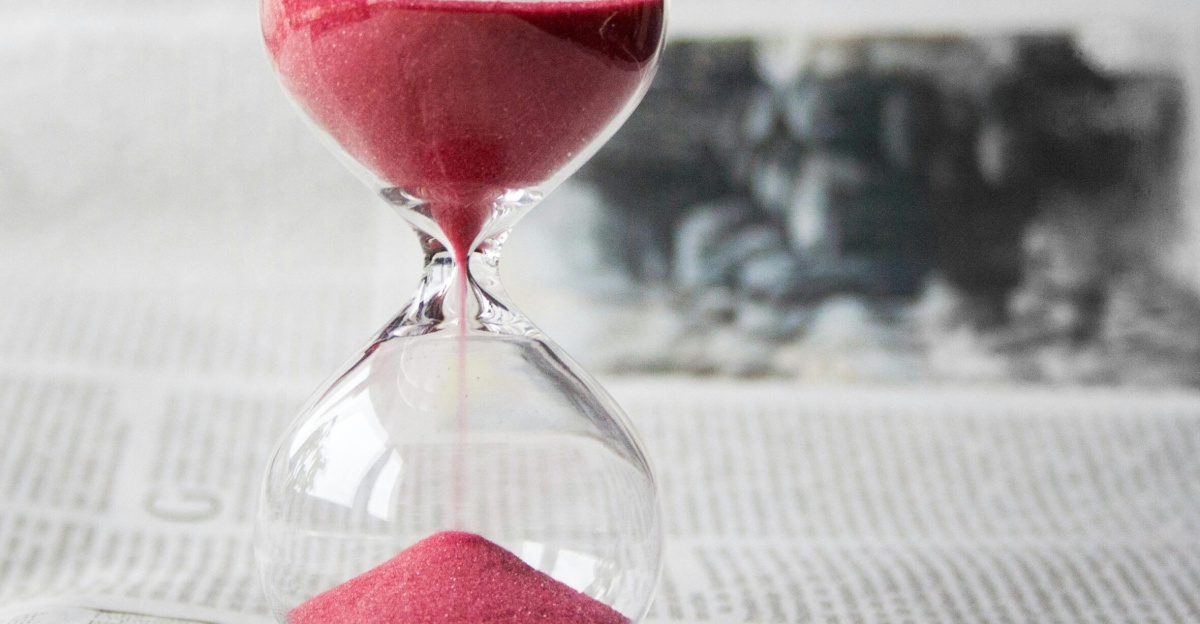 Close-up of a transparent hourglass with pink sand flowing, placed on a newspaper background.