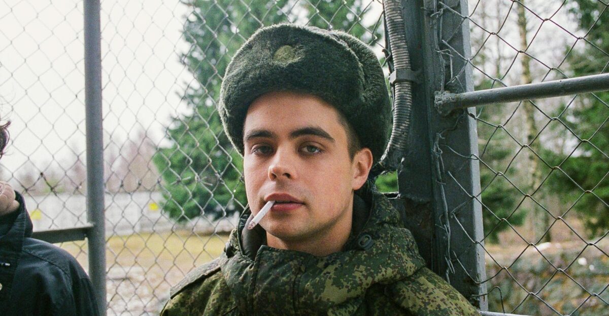 A young soldier in camouflage uniform smokes a cigarette by a chain-link fence in a winter setting
