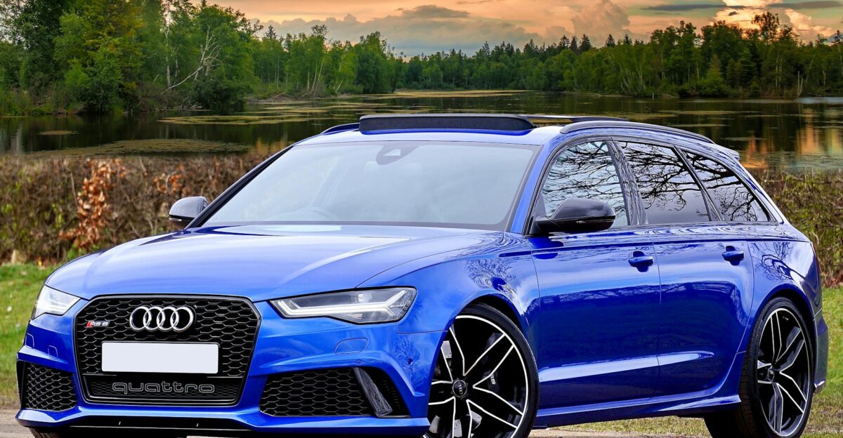 A modern blue Audi RS6 parked by a lake with scenic forest and sky backdrop