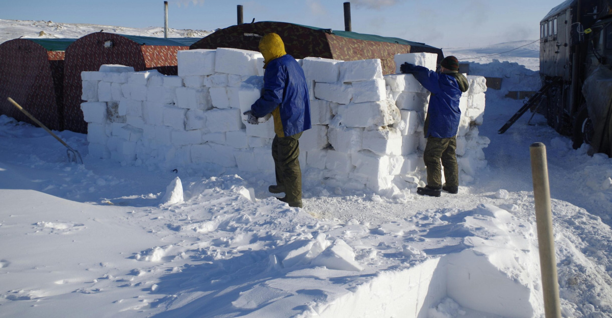 Two people stacking snow blocks to build a wall in a snowy landscape.