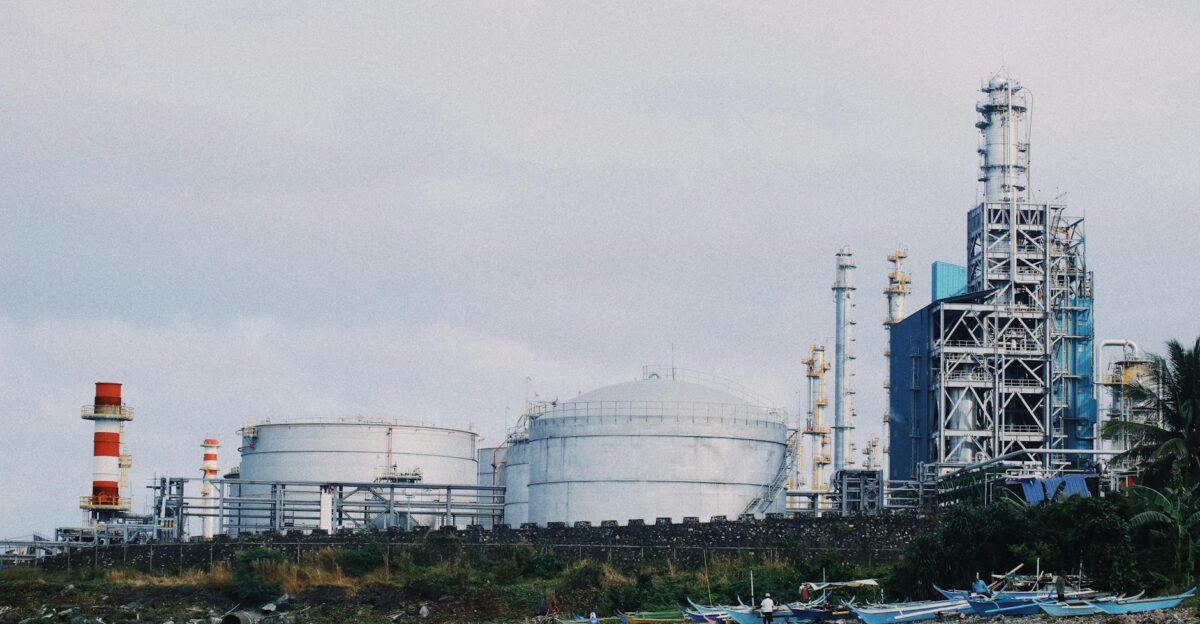 A coastal industrial petrochemical plant with storage tanks and factory structures against a cloudy sky