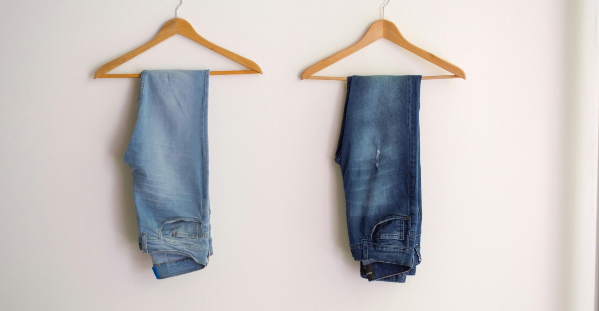 Two pairs of blue denim jeans neatly hanging on wooden hangers against a plain wall.
