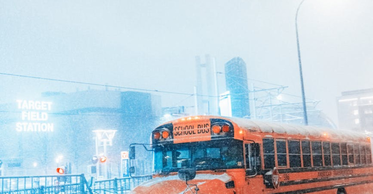 A school bus navigates a snow-covered road amidst city buildings during a heavy snowfall
