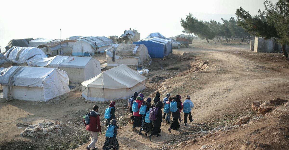 Children carrying backpacks walk through a rural refugee camp under a clear sky