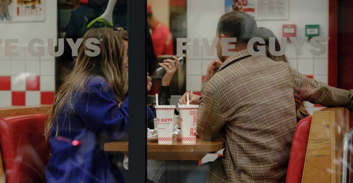 A group of friends share a meal and conversation at Five Guys visible through the window