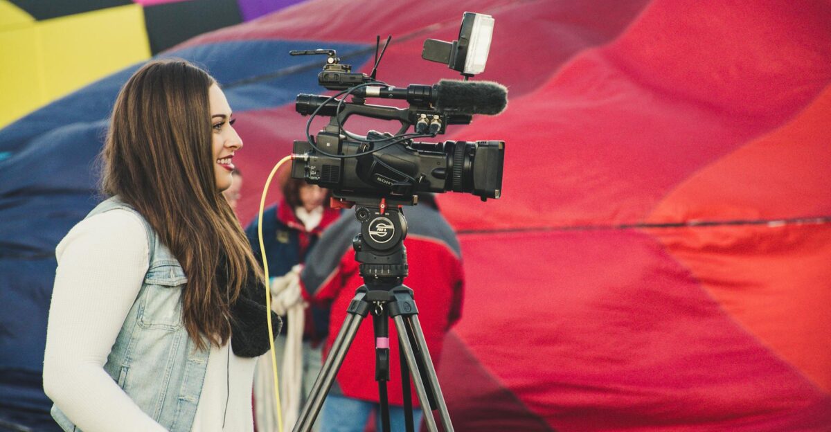 A female videographer operates a camera before a colorful hot air balloon backdrop