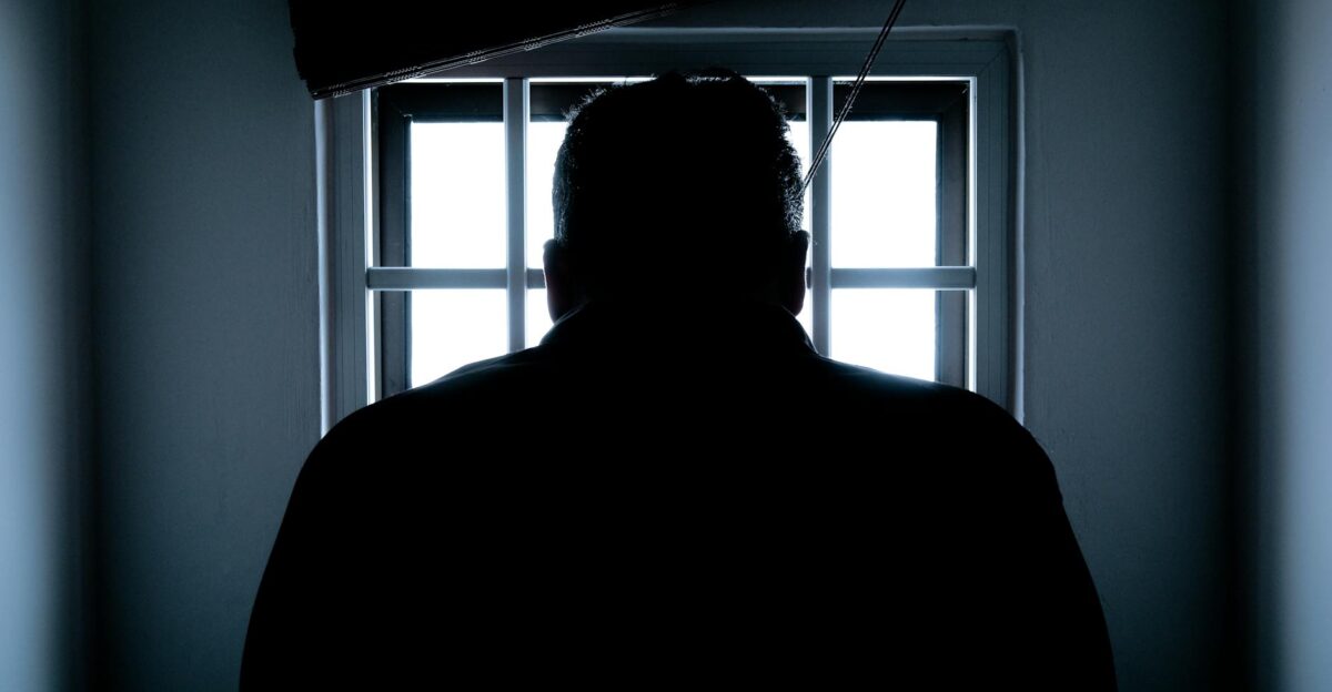 Dark silhouette of a man in a prison cell backlit by window bars