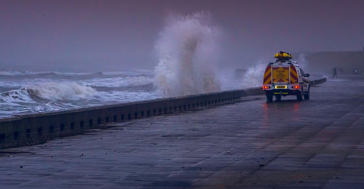 A dramatic coastal scene with waves crashing against a seawall and a vehicle on a stormy day.