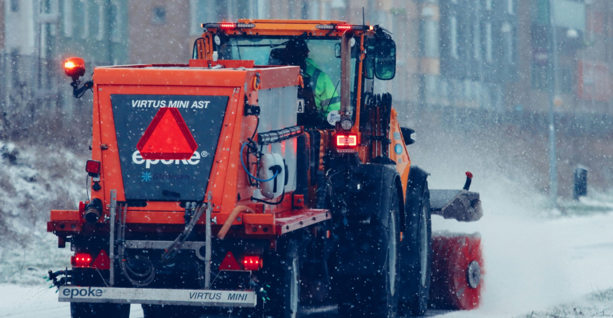 Orange snowplow in action clearing a snow-covered street during a winter snowfall.
