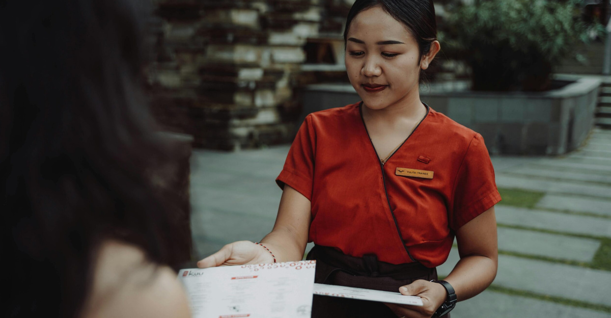 A waitress in a red uniform handing a menu to a guest at an outdoor restaurant.