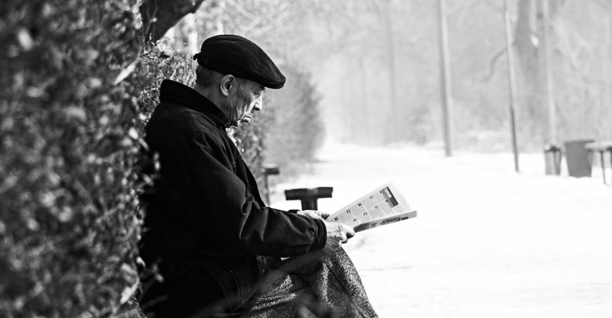 A senior man reads a newspaper on a snowy bench in a tranquil winter park